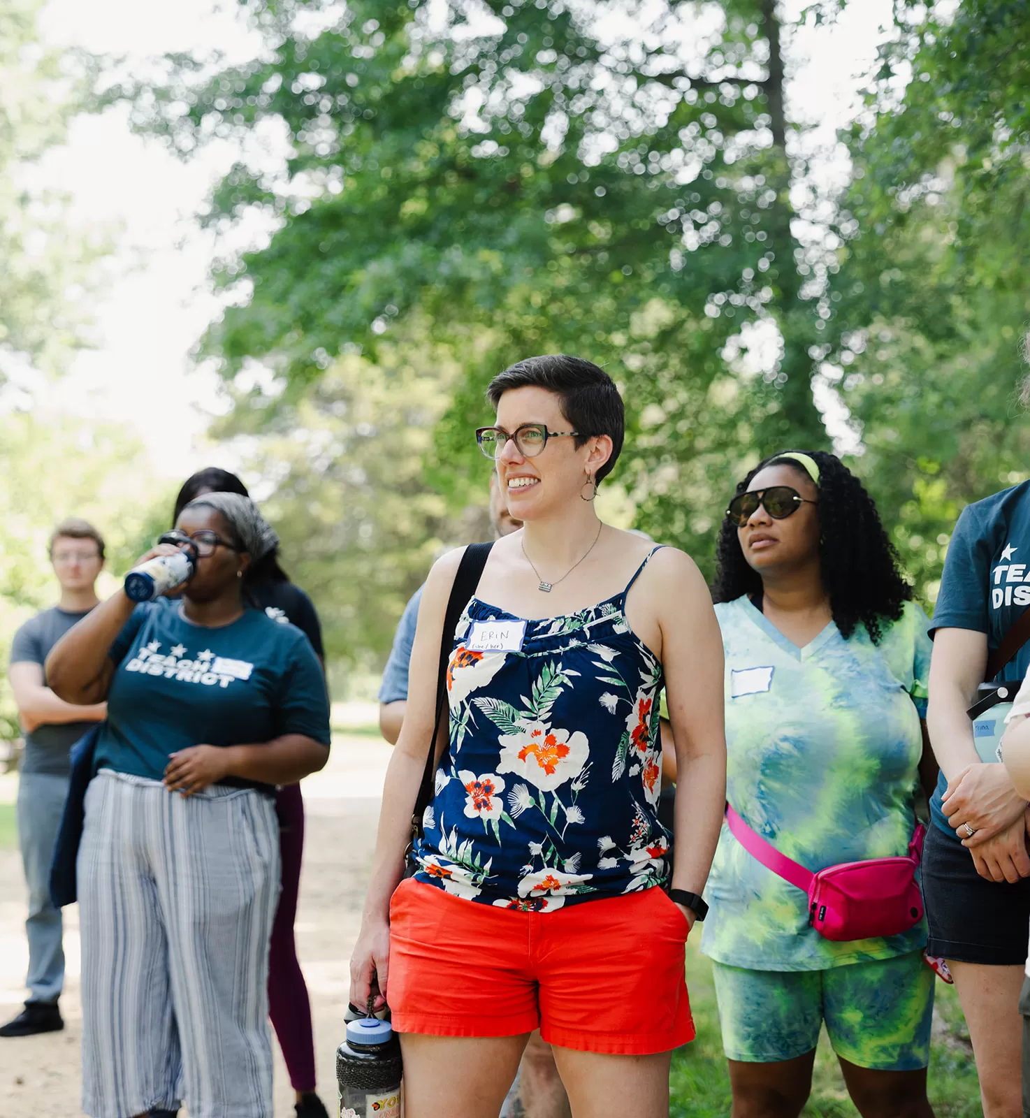 A group of people stands outdoors on a sunny day, surrounded by trees. In the foreground, a person wearing glasses, a floral tank top, and red shorts smiles while holding a water bottle. Others in the group wear casual clothing, including one person in a green tie-dye outfit with a pink waist bag and another sipping from a water bottle. Everyone appears to be engaged in an outdoor activity or event.