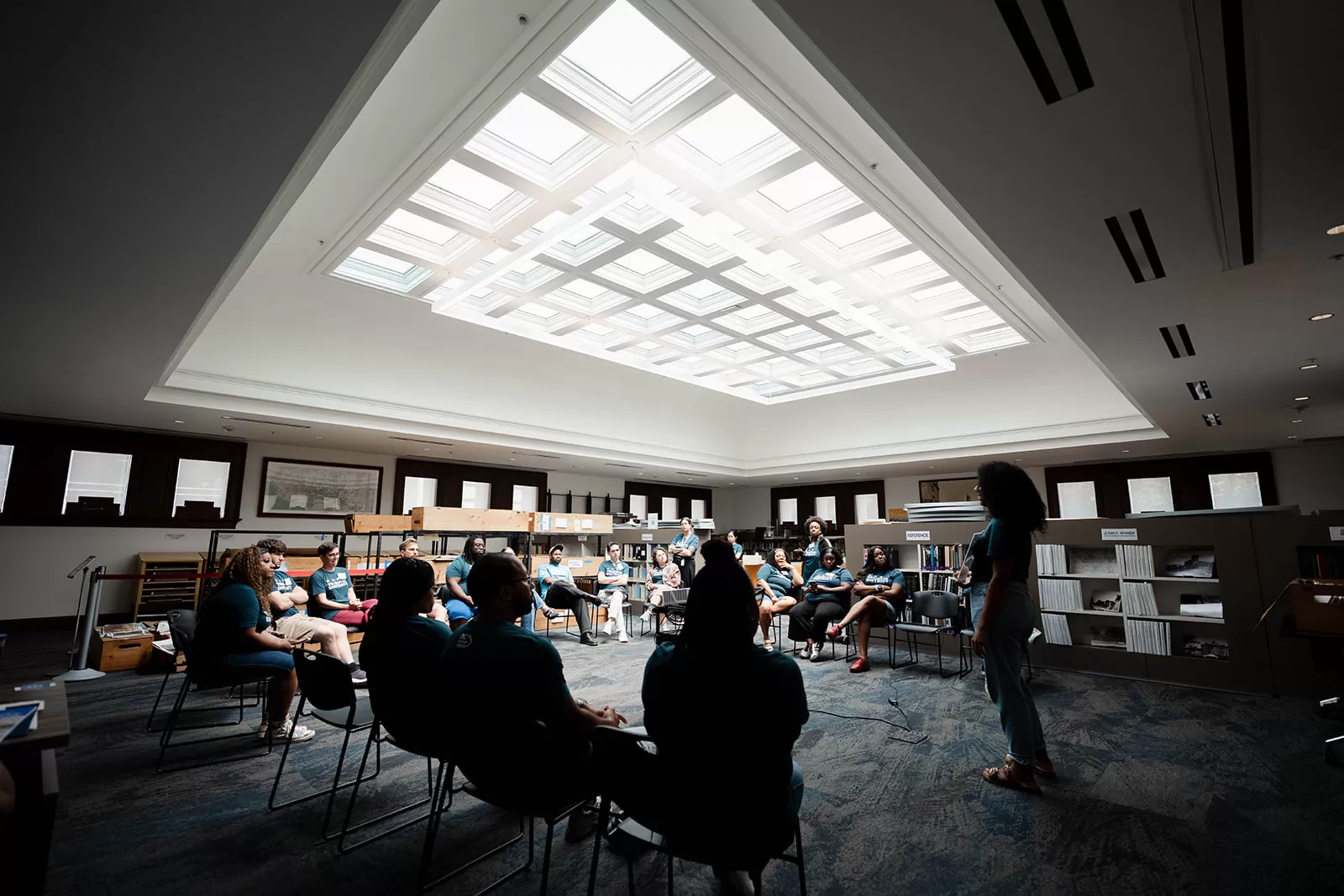 A group of people seated in a circle inside a bright, modern library or archive room, listening to a speaker standing in the foreground. Natural light pours in from a large skylight above, illuminating the space. Shelving and boxes line the walls, indicating a research or educational setting.