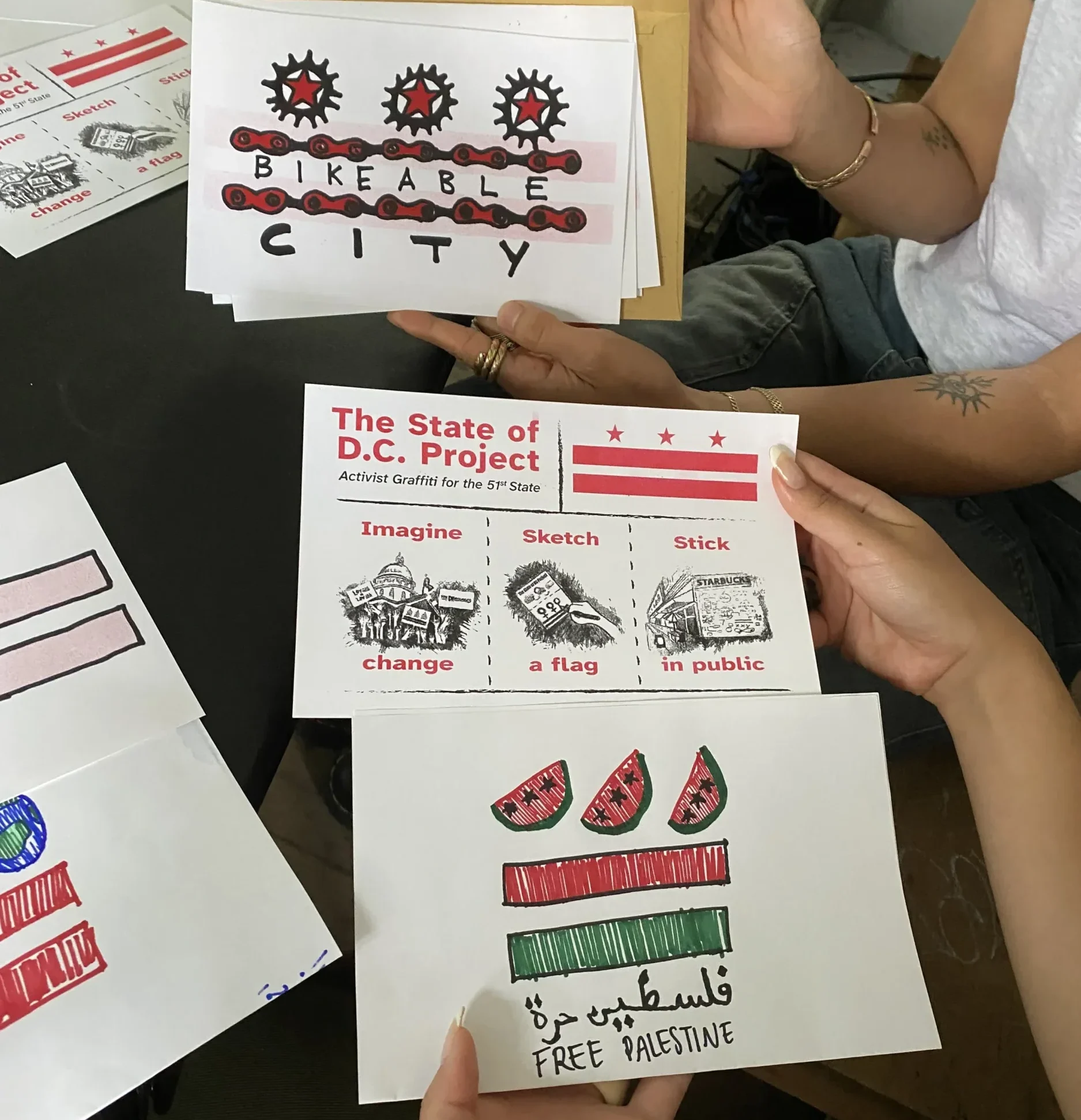 Three people sit around a table holding handmade postcard-sized protest artworks. One card reads “Bikeable City” with drawings of bike gears and a red chain. Another is titled “The State of D.C. Project: Activist Graffiti for the 51st State” with small illustrations and the words “Imagine change, Sketch a flag, Stick in public.” A third card shows red and green bars with watermelon slices above and the text “Free Palestine” in English and Arabic.