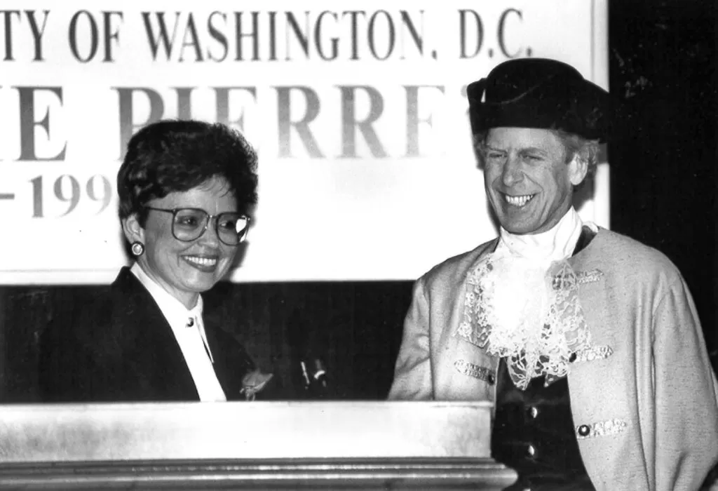 A woman in a suit and a man dressed in 18th-century colonial attire smile at a podium during a public event in Washington, DC. A large sign in the background reads “PIERRE” and includes a partial date, suggesting a historical or commemorative occasion.